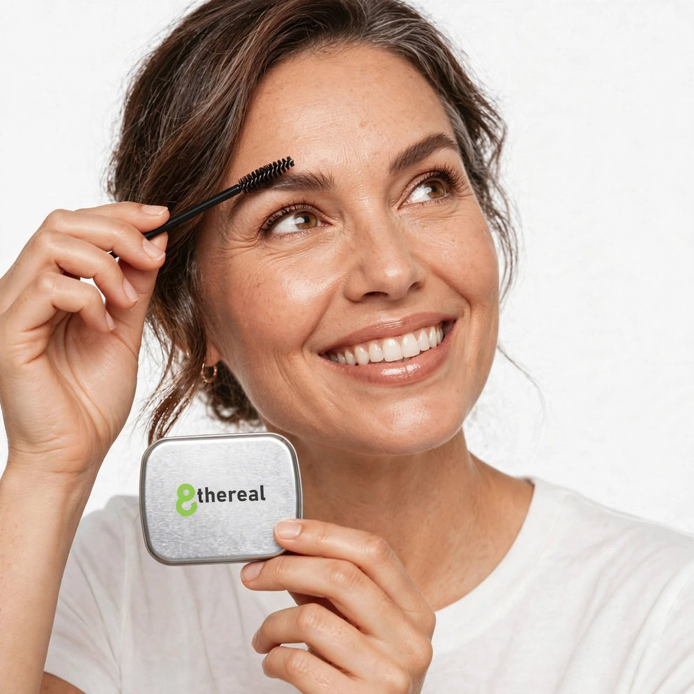 A smiling woman applying 8thereal clear brow soap with a spoolie to lift and shape her natural eyebrows.