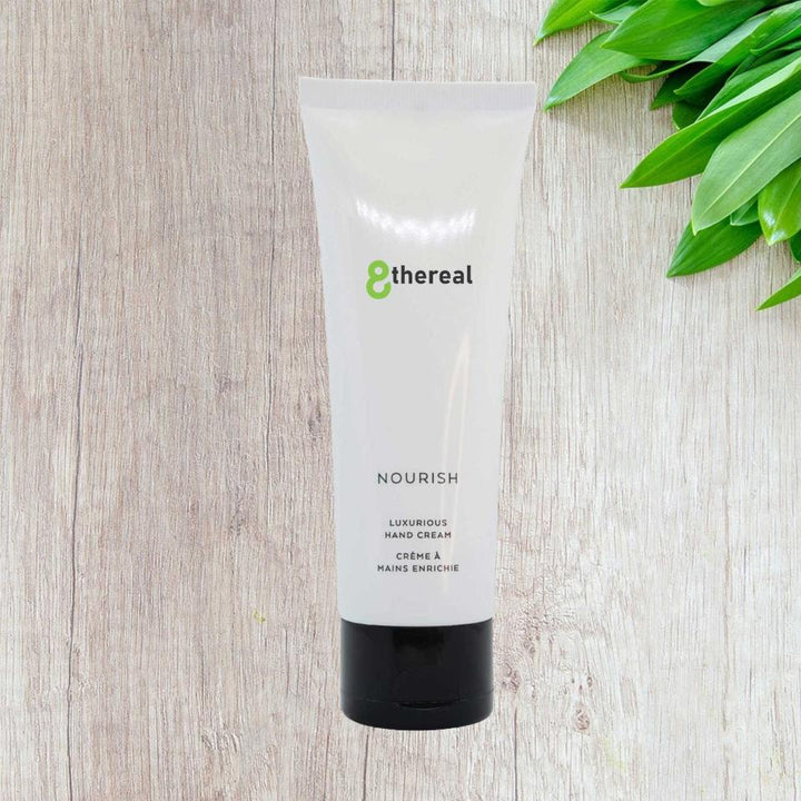 8thereal Restorative Nourish Hand Cream in a white squeeze tube on a wooden surface with green leaves.